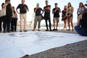 En primer plano en el piso y en el fondo, de pie, alumnos Arquitectura UDD detenidamente el mapa